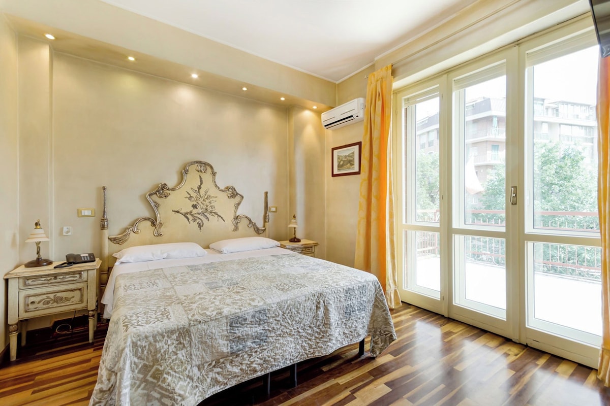 A bedroom with a large bed, wooden flooring, an ornate headboard, bedside tables with lamps, and a large window with curtains.