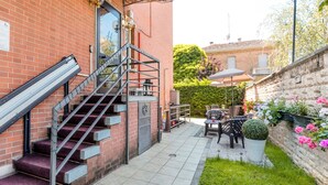 A brick building with a staircase leading to a door, a patio area with a table and chairs, and a garden with flowers and a hedge. - Hotel Original (Turin)