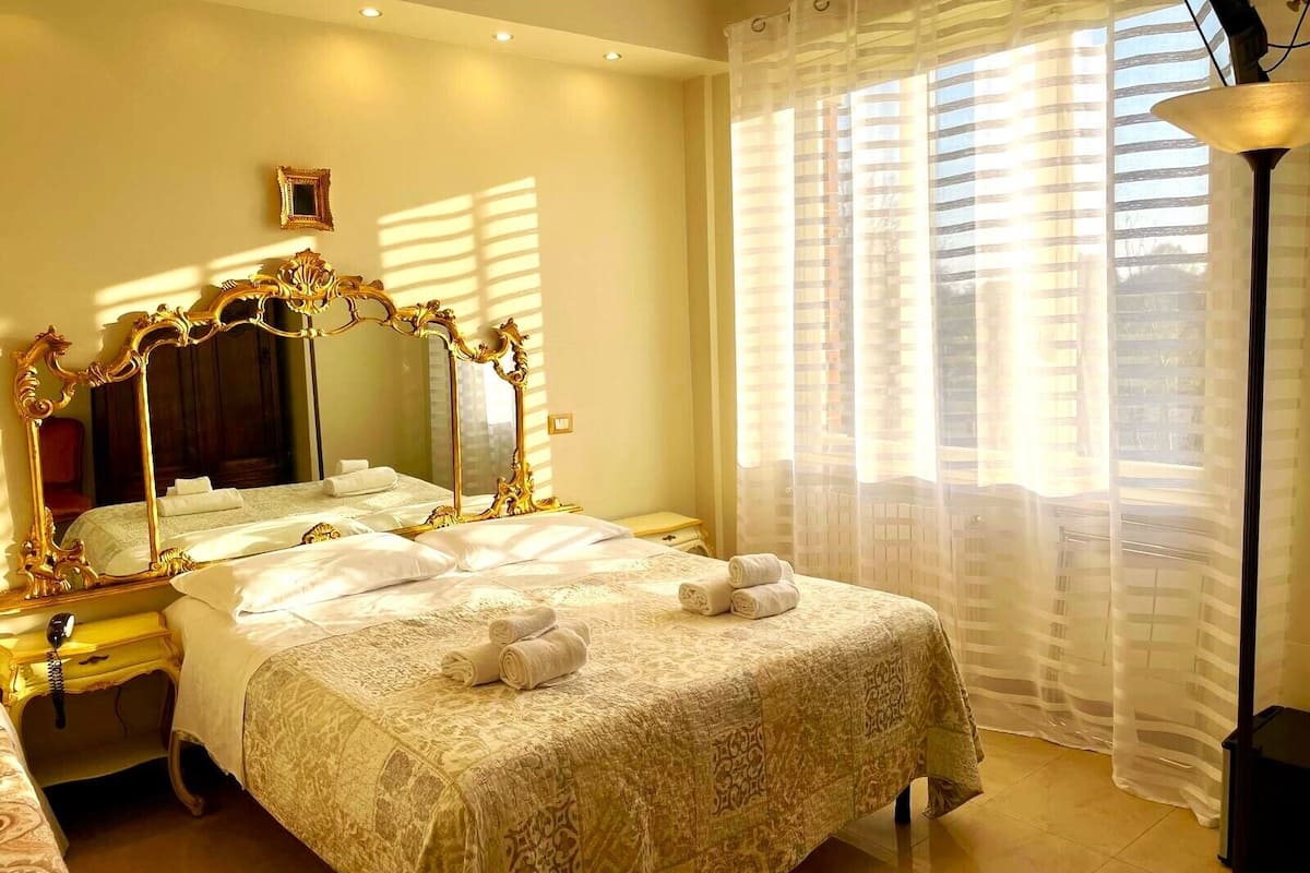 A hotel room with a large bed, a bedside table with a lamp, and a window with sheer curtains.
