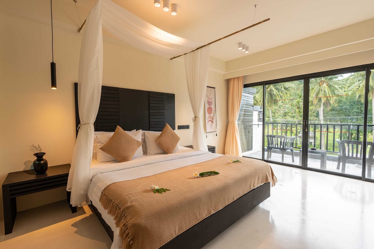 A modern bedroom with a large bed, a nightstand, a bedside table, and a view of the outdoors through large windows.