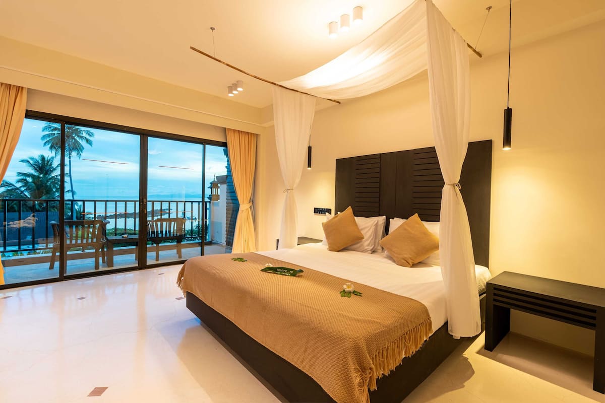 A hotel room with a large bed, a nightstand, and a view of palm trees through a balcony.