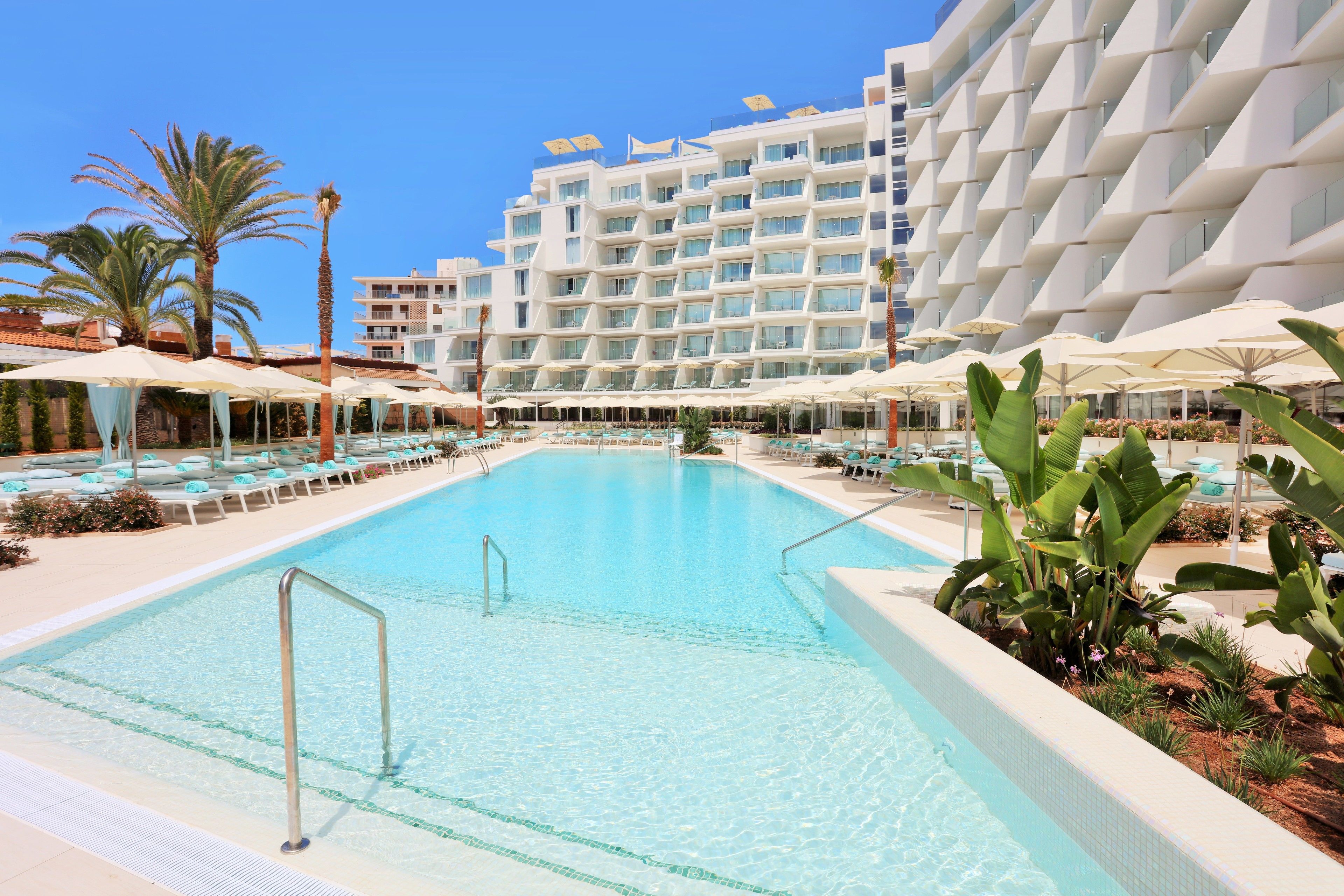 A swimming pool with lounge chairs and umbrellas, surrounded by a hotel building with multiple floors and balconies.