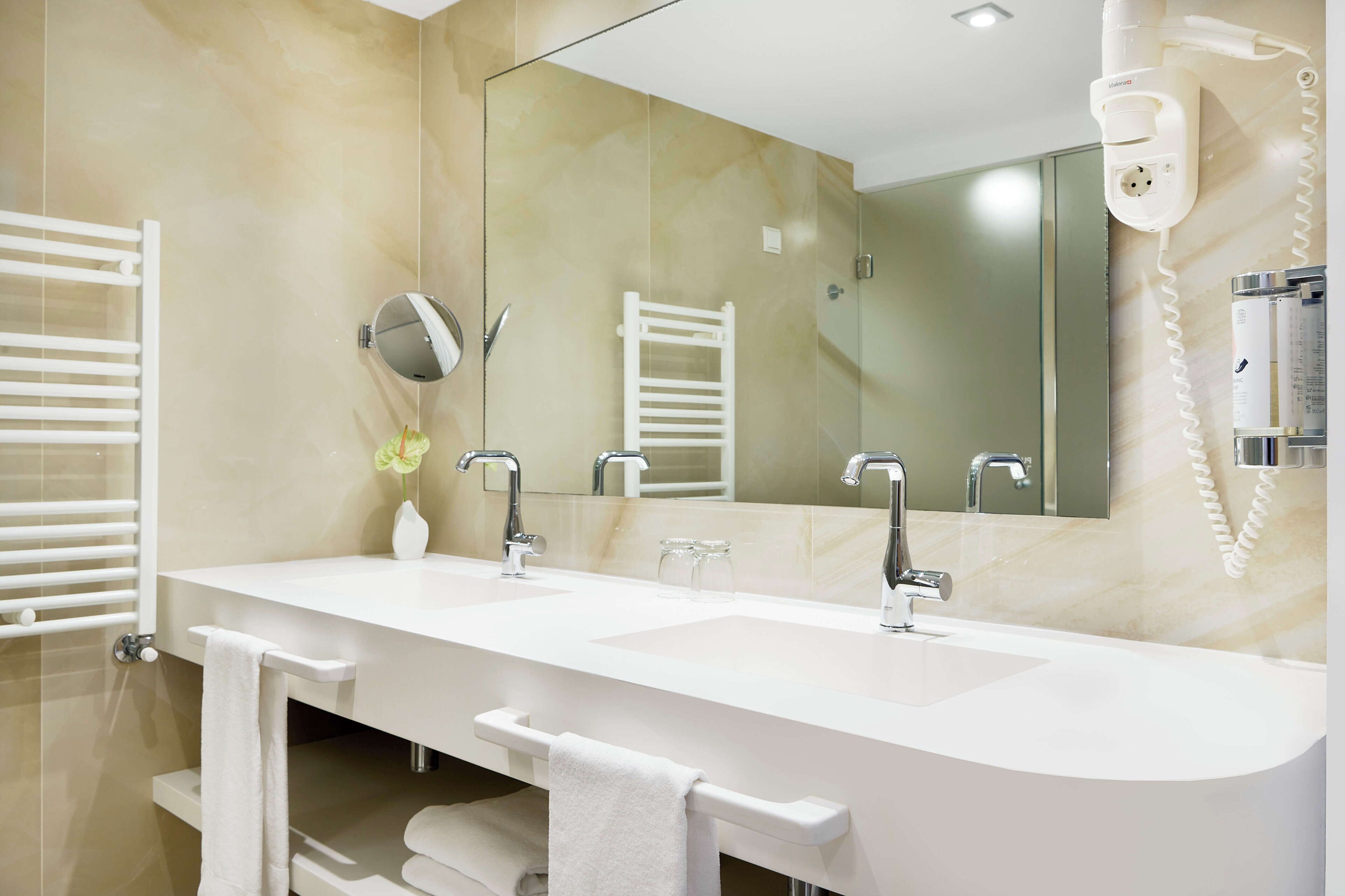 Une salle de bain moderne avec un double vasque, un grand miroir, un porte-serviettes et un sèche-cheveux.
