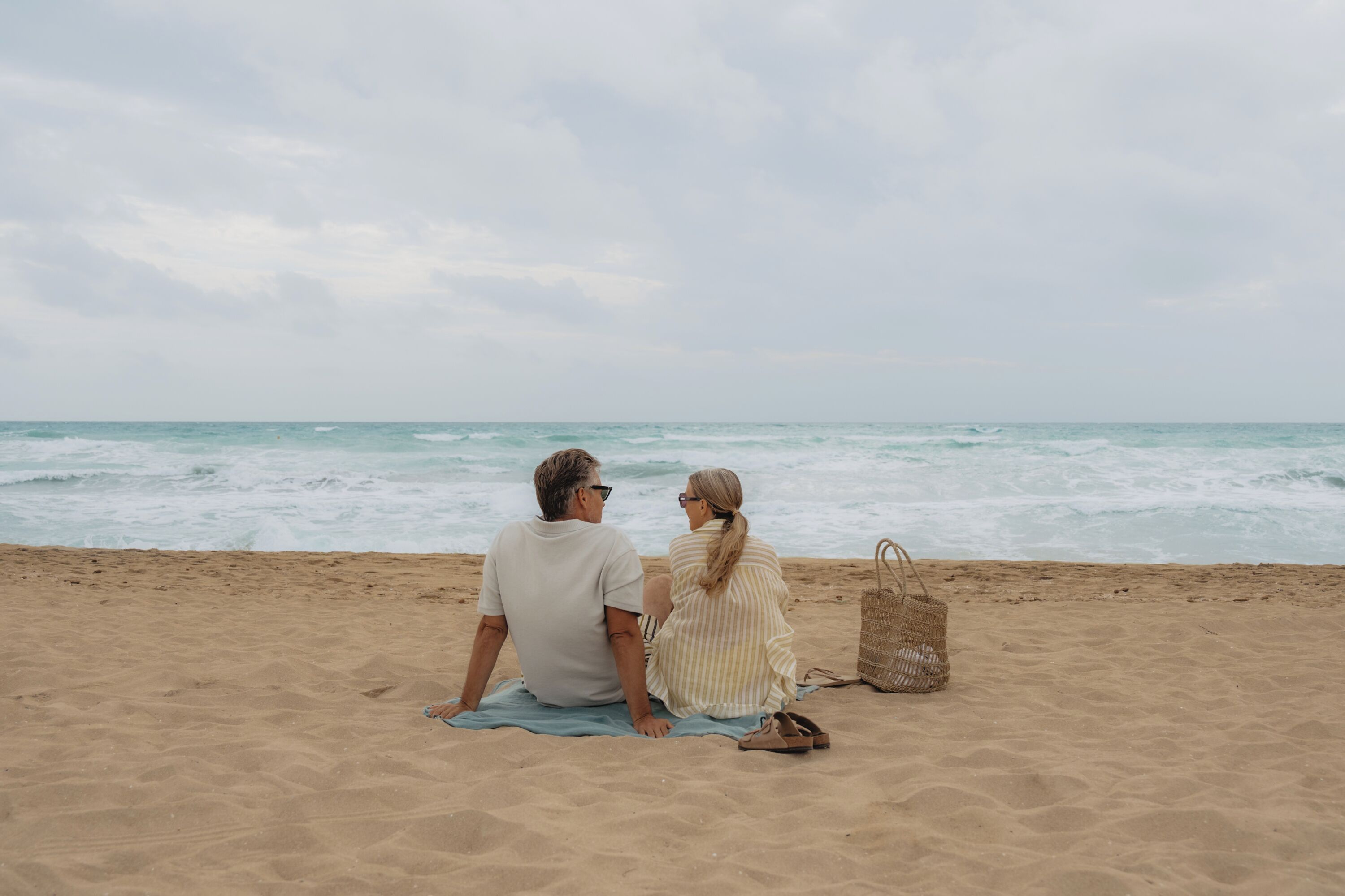 A couple sitting on a beach towel facing the ocean.