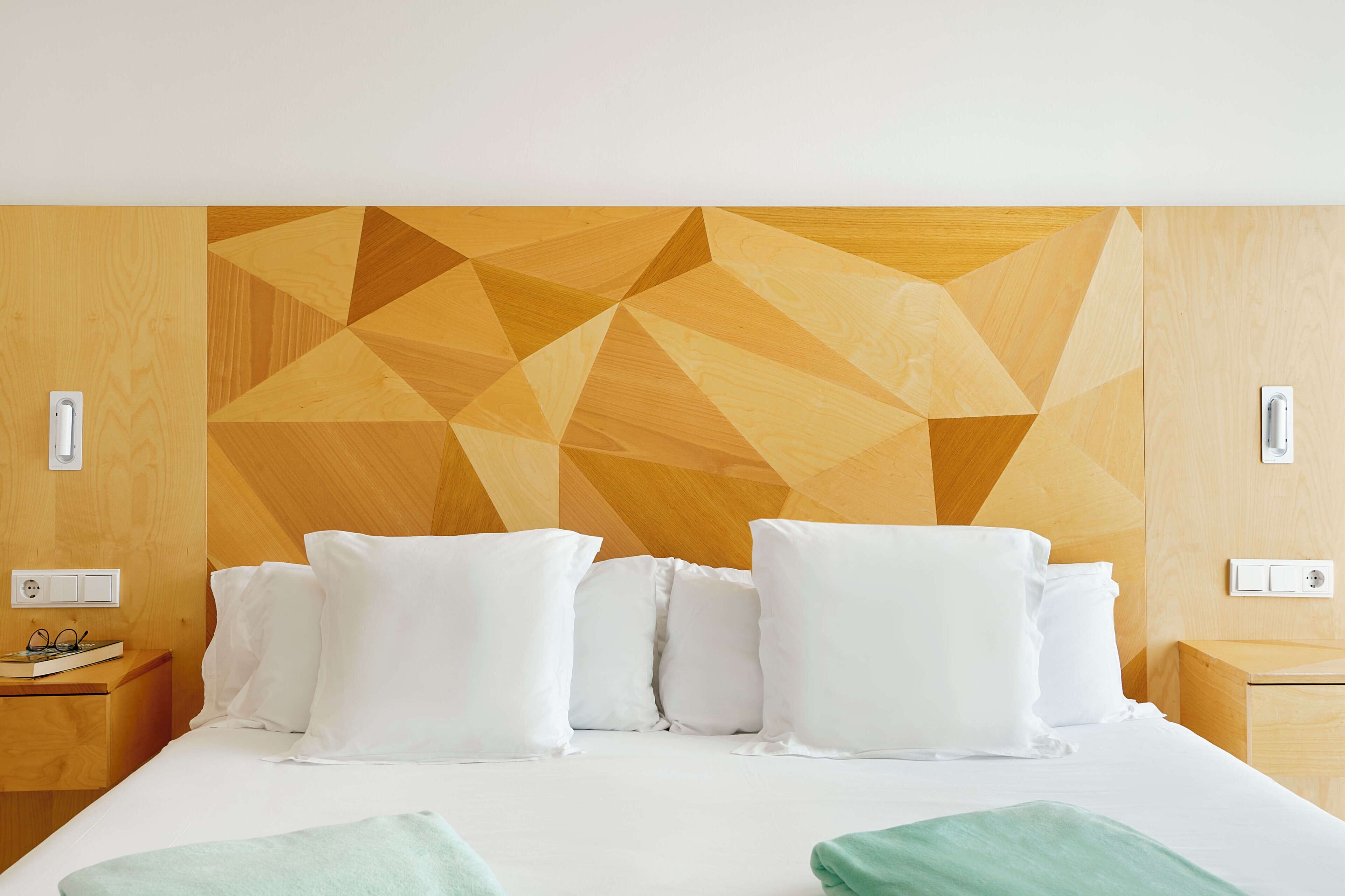 A hotel room with a geometric-patterned headboard, a neatly made bed with white linens, and a bedside table with a book and glasses.