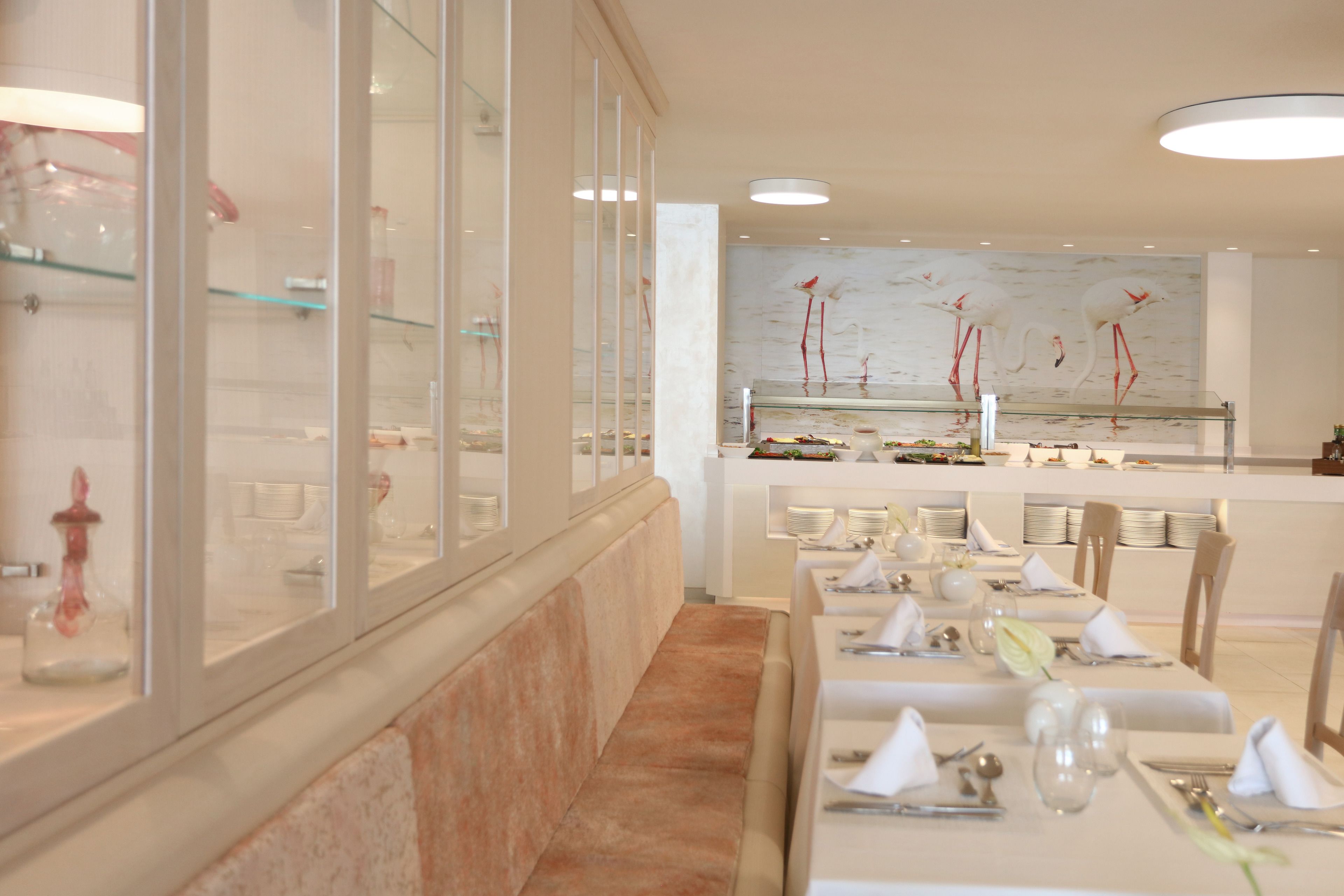 En restaurant med glasfacade, pænt anrettede borde med hvide duge og en marmorvæg med et fuglemotiv.
