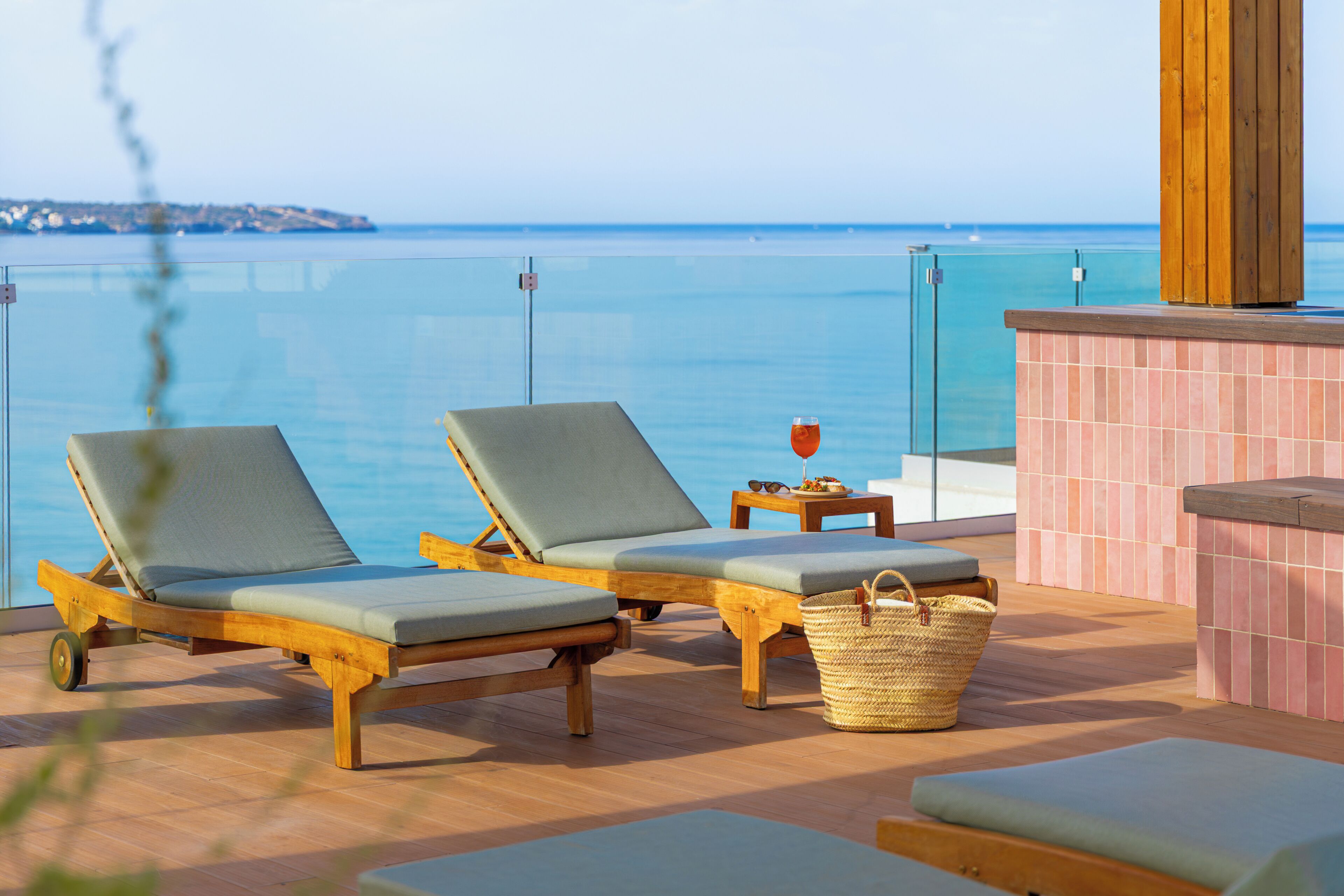 A sunlit outdoor lounge area with two wooden lounge chairs, a small table with a drink, and a wicker basket.