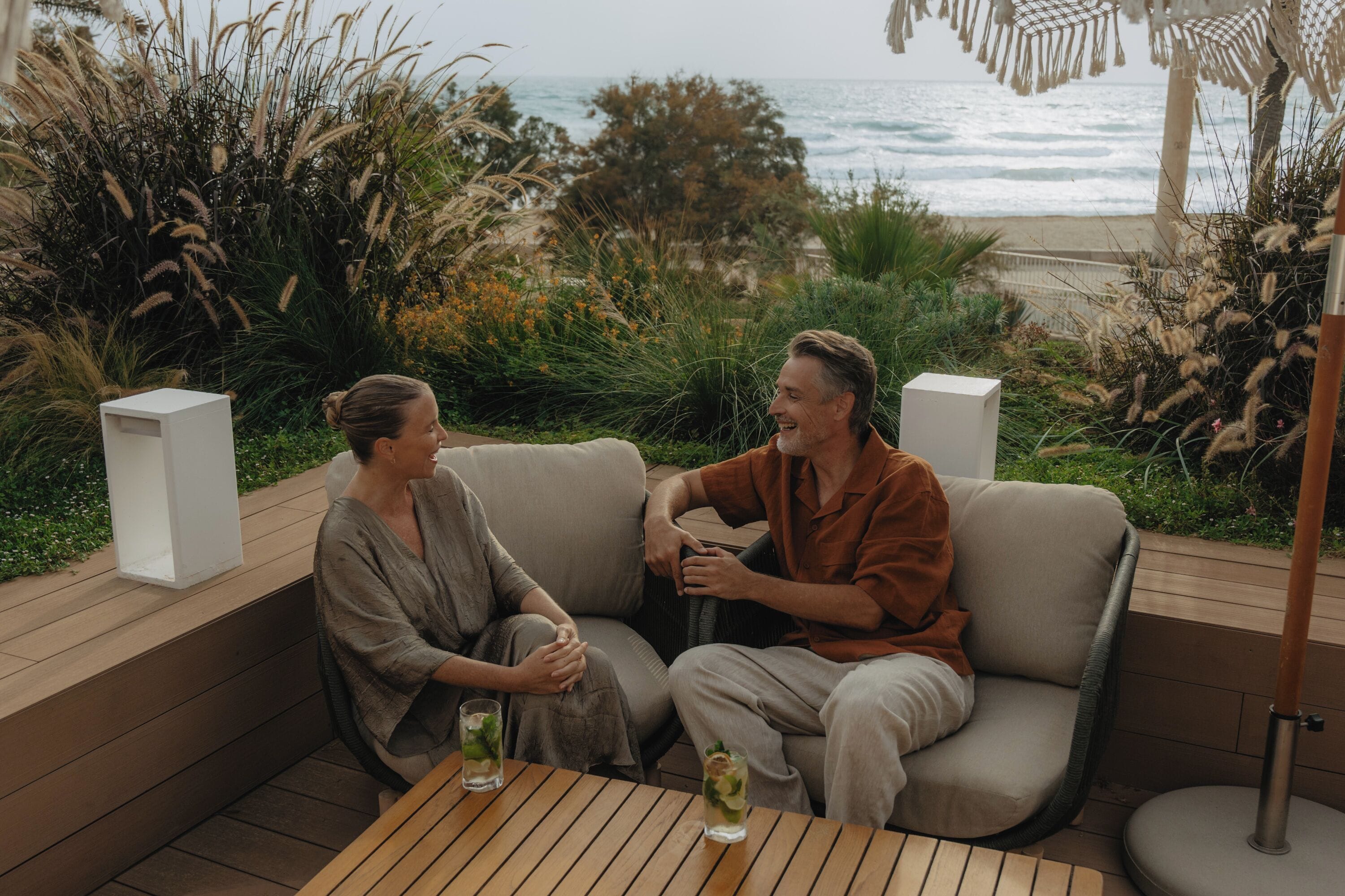 Un couple est assis sur un canapé confortable installé à l’extérieur, devant une table en bois, et profite d’une vue imprenable sur la plage et l’océan.