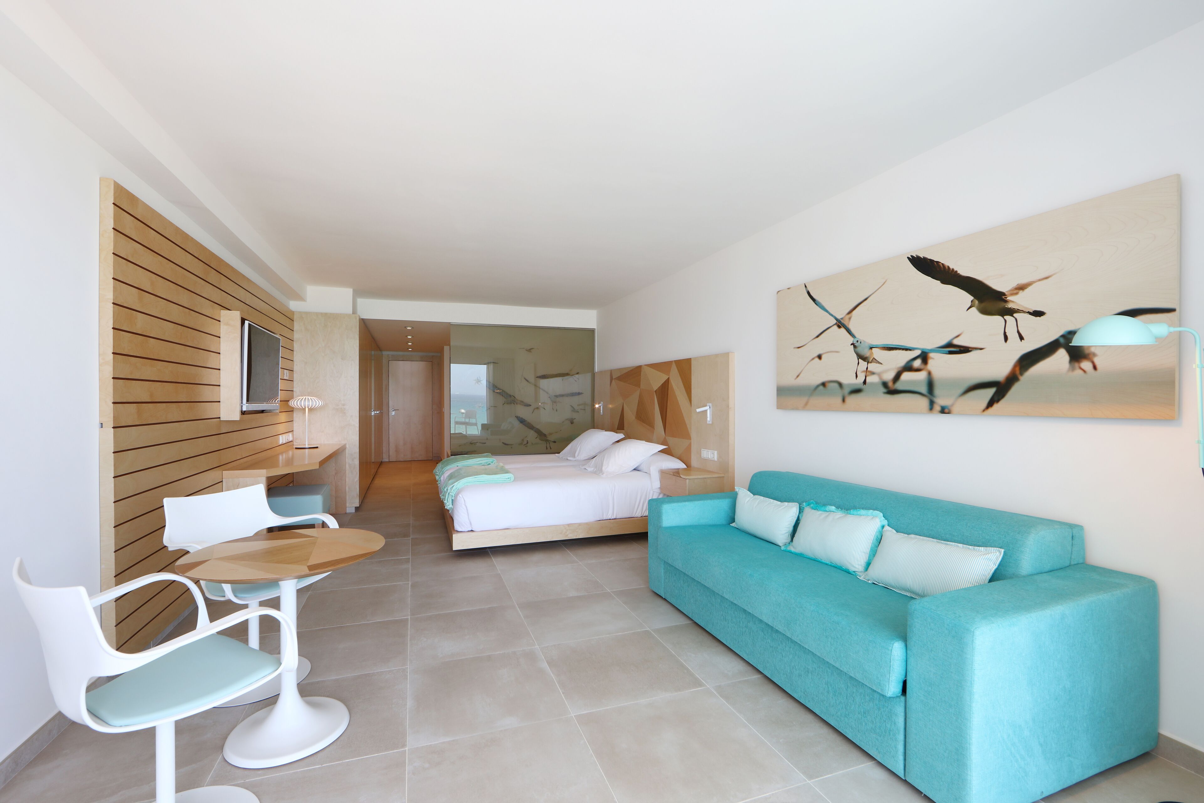 Una habitación de hotel moderna con una cama, un sofá color turquesa, una mesa de comedor con sillas y un cuadro de pájaros en la pared.