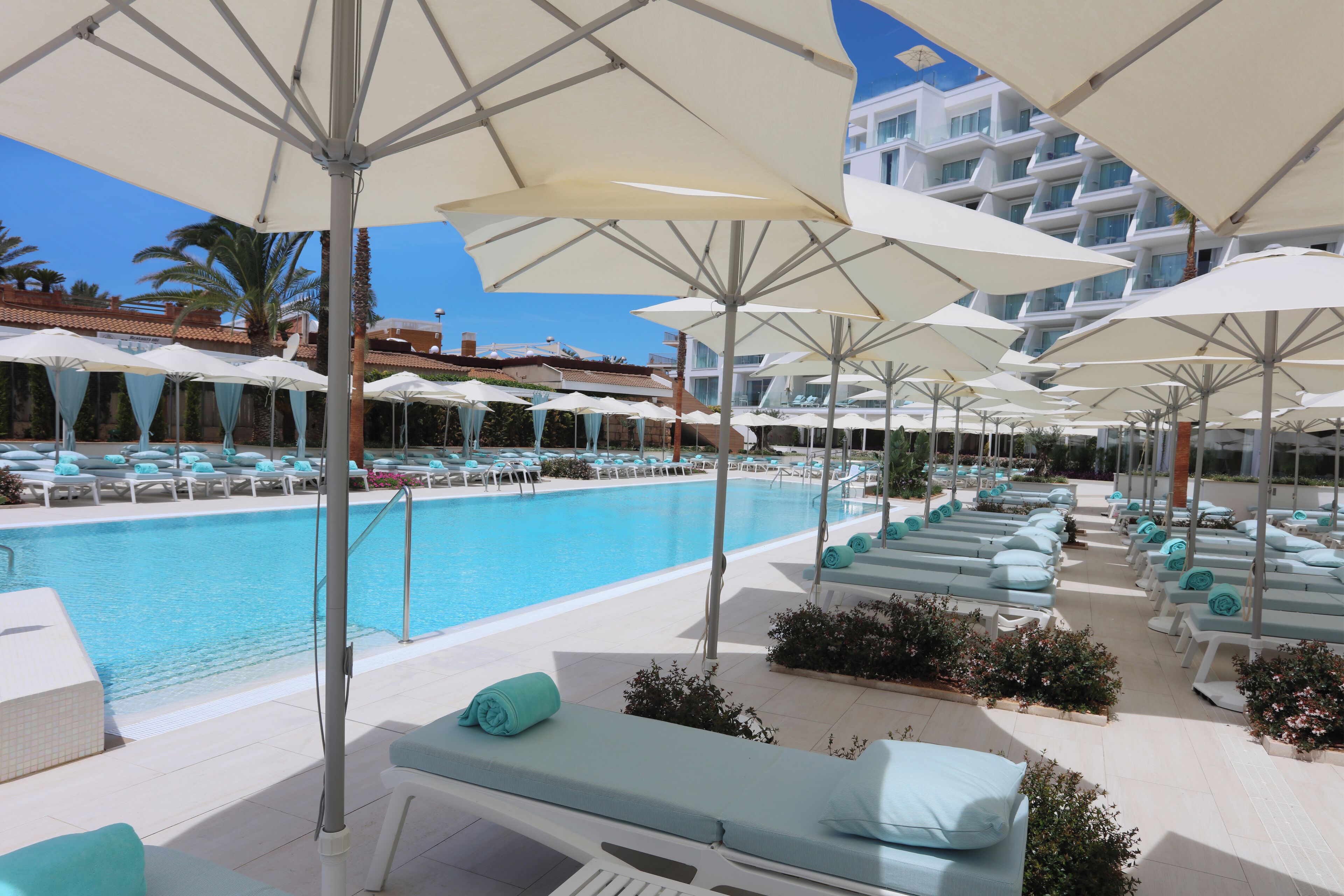 A poolside area with multiple lounge chairs and umbrellas, a swimming pool, and a building with balconies in the background.