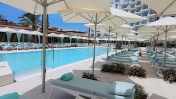 Zona de piscina amb diverses gandules i ombrel·les, piscina i edifici al fons amb balcons.