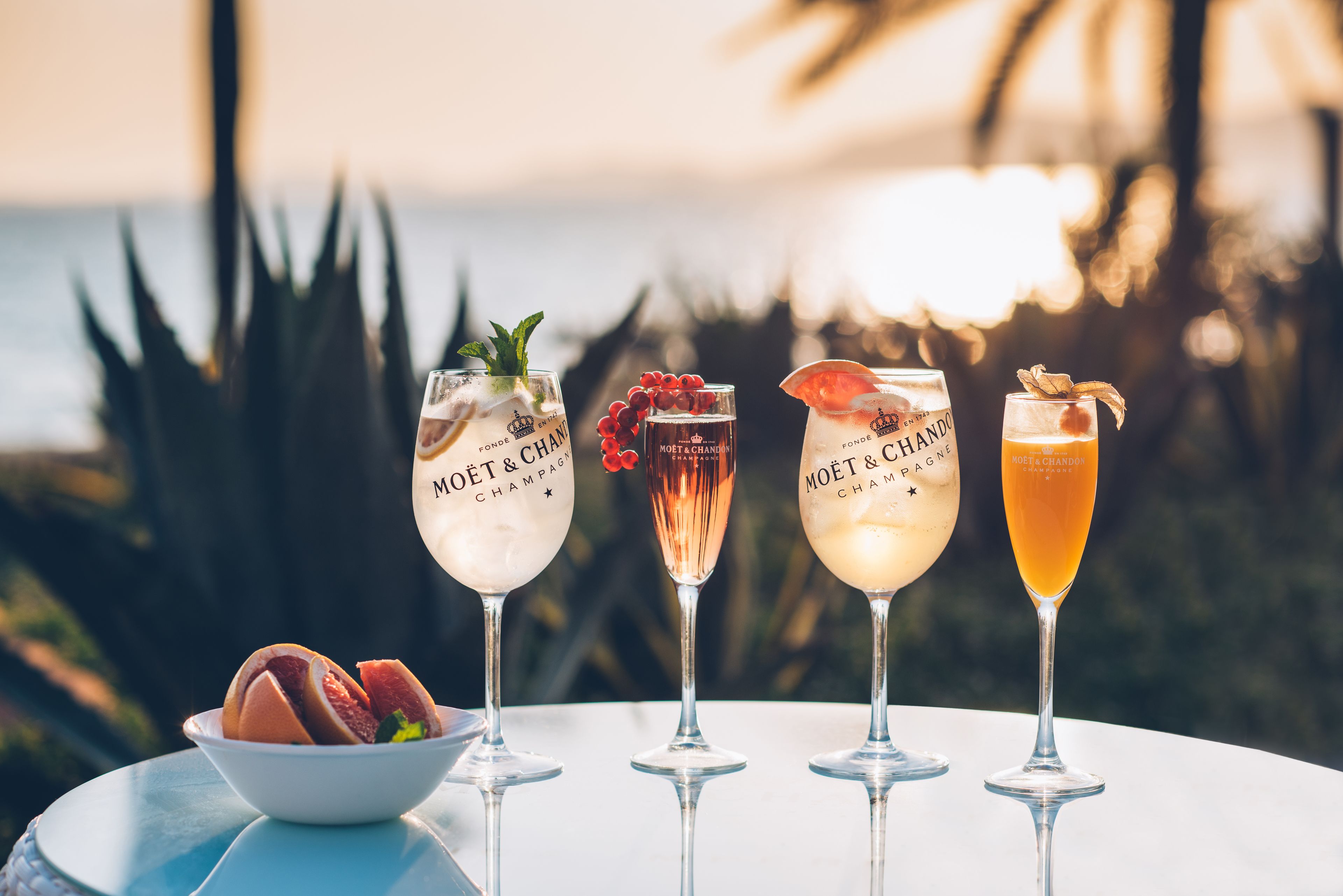 Four glasses of Moët & Chandon champagne on a table with a bowl of fruit and a view of the ocean.