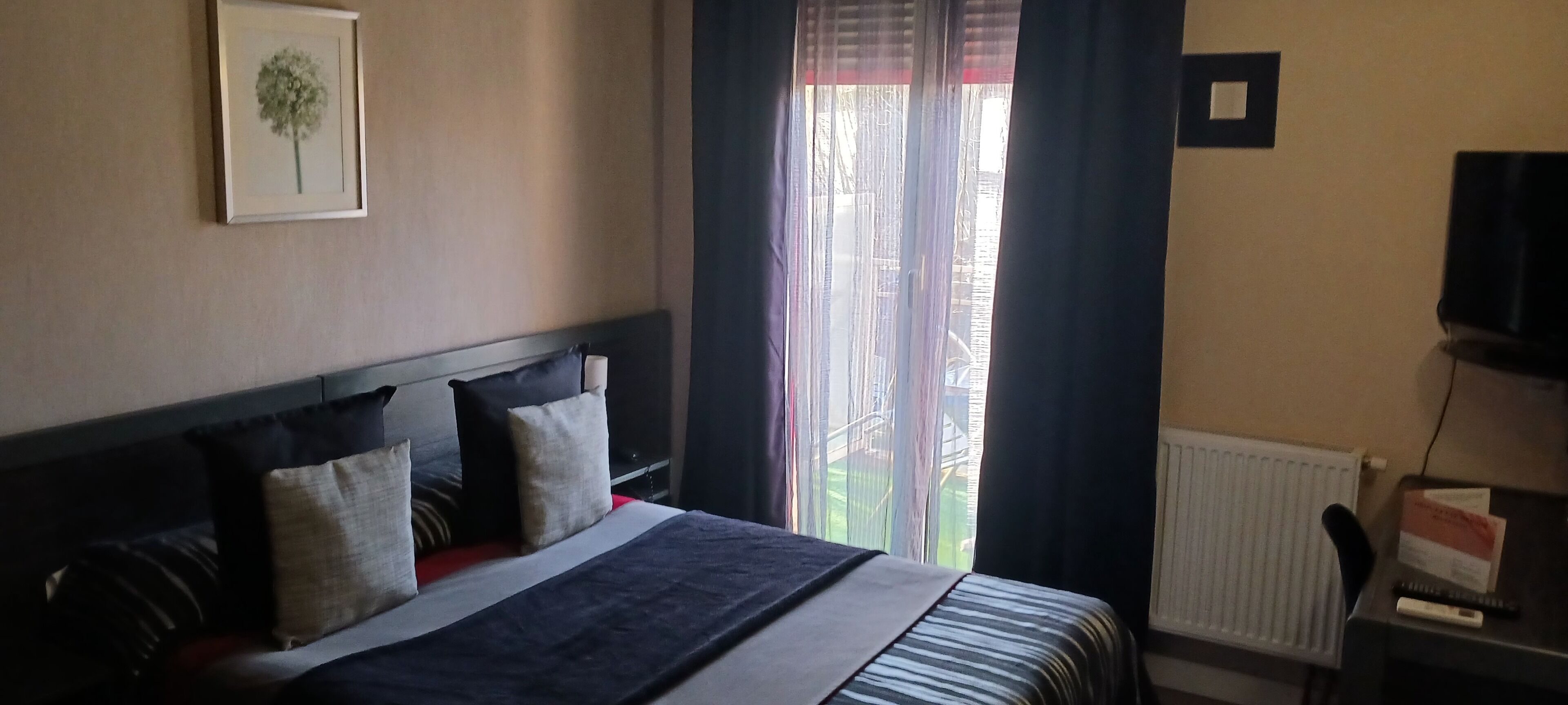 Habitació d'hotel amb un llit, un quadre amb marc a la paret, una finestra amb cortines, un radiador i una televisió sobre un suport.