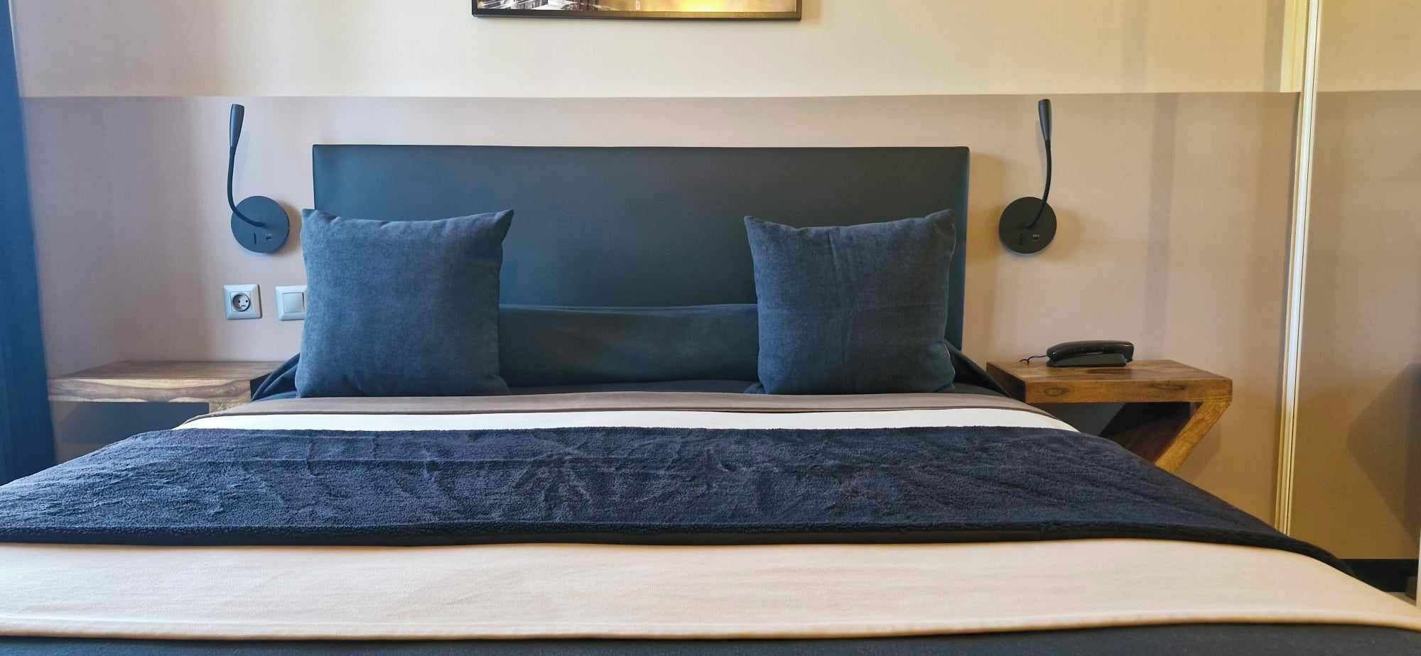 Una cama con cabecera oscura, dos almohadas, una colcha oscura y una mesita de noche de madera con una lámpara.