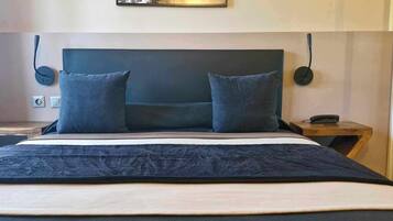 Una cama con cabecera oscura, dos almohadas, una colcha oscura y una mesita de noche de madera con una lámpara.