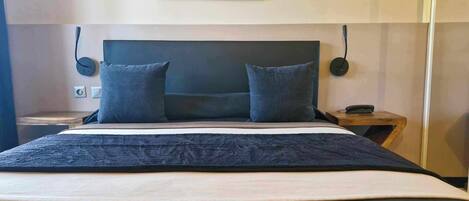 Una cama con cabecera oscura, dos almohadas, una colcha oscura y una mesita de noche de madera con una lámpara.