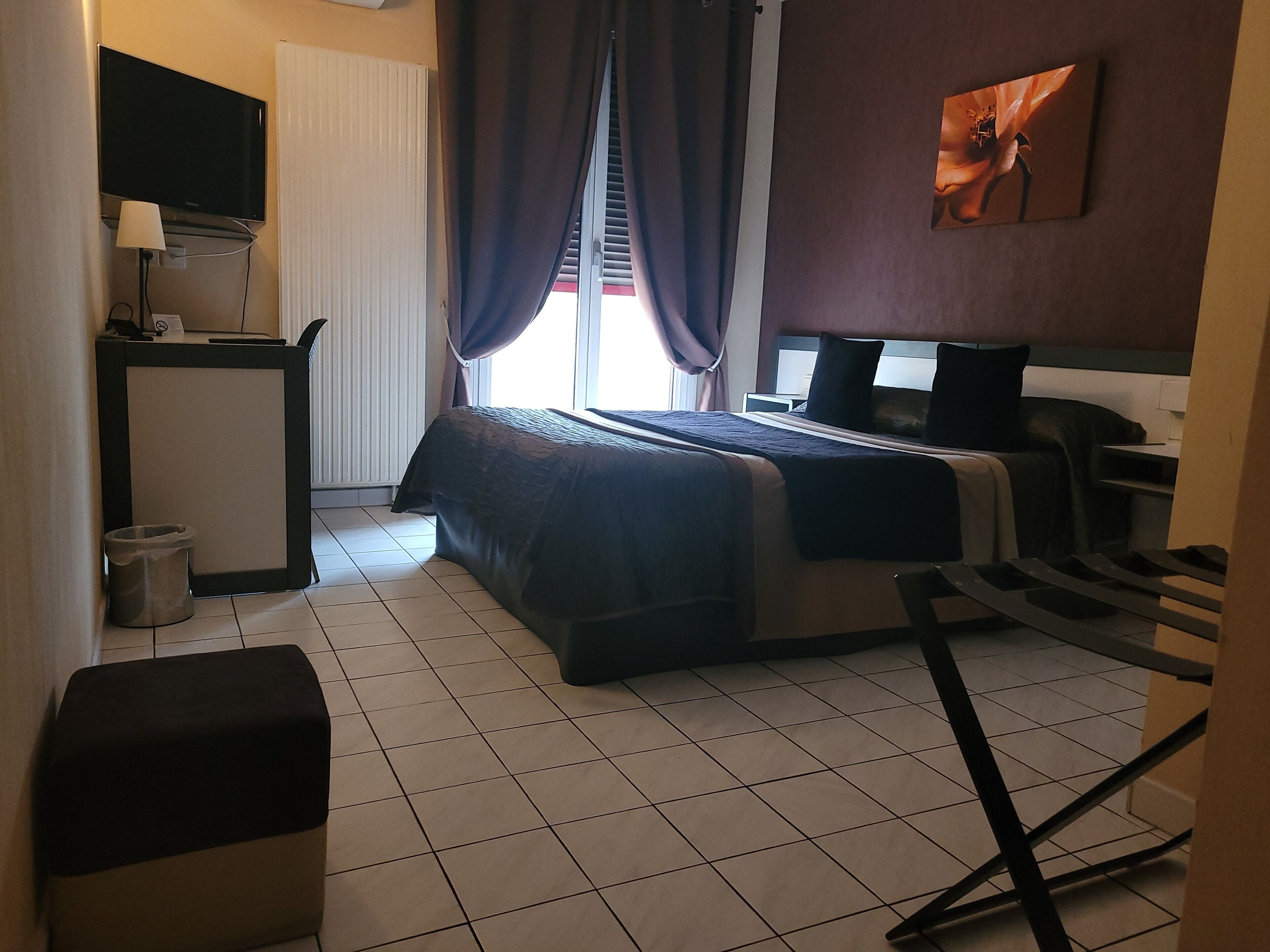 A hotel room with a bed, a desk with a lamp, a TV, and a painting on the wall.
