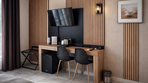 A modern hotel room with a wooden desk, two black chairs, a flat-screen TV mounted on the wall, a small trash bin, and a framed picture on the wall.