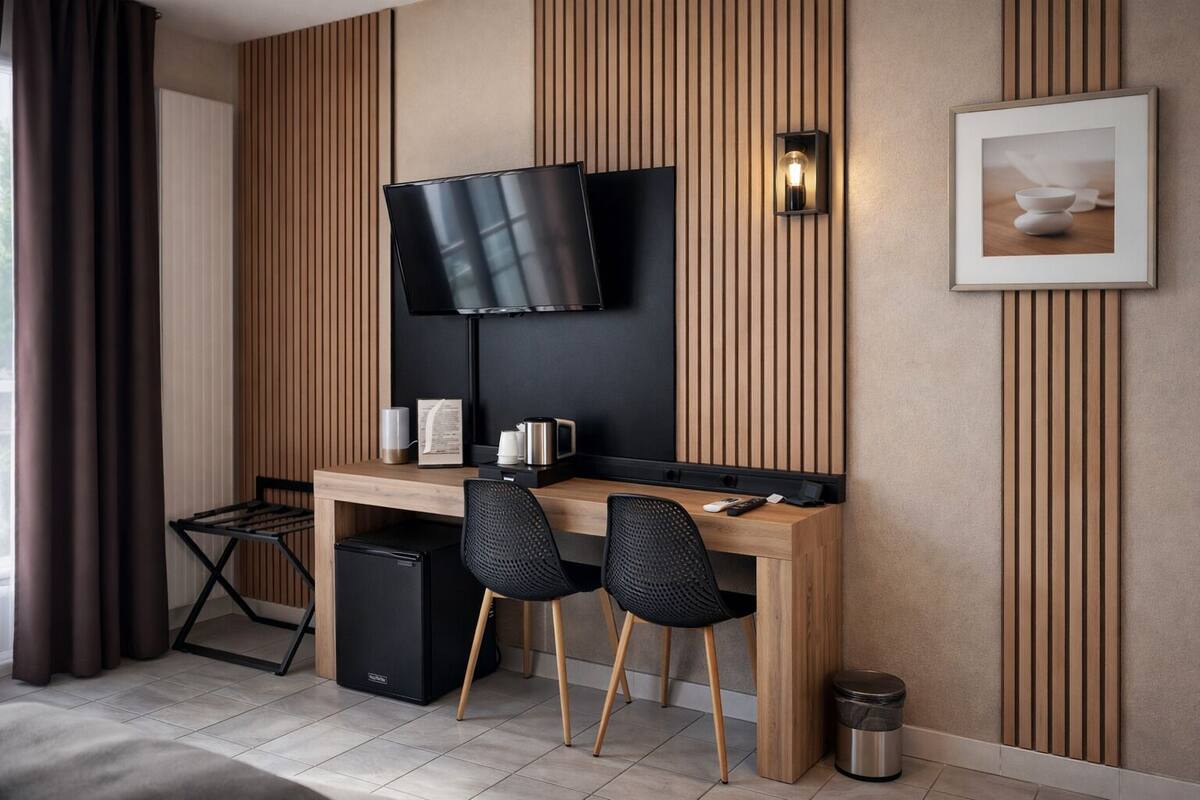 Une chambre d’hôtel moderne avec un bureau en bois, deux chaises noires, un téléviseur à écran plat fixé au mur, une petite poubelle et un tableau encadré accroché au mur.
