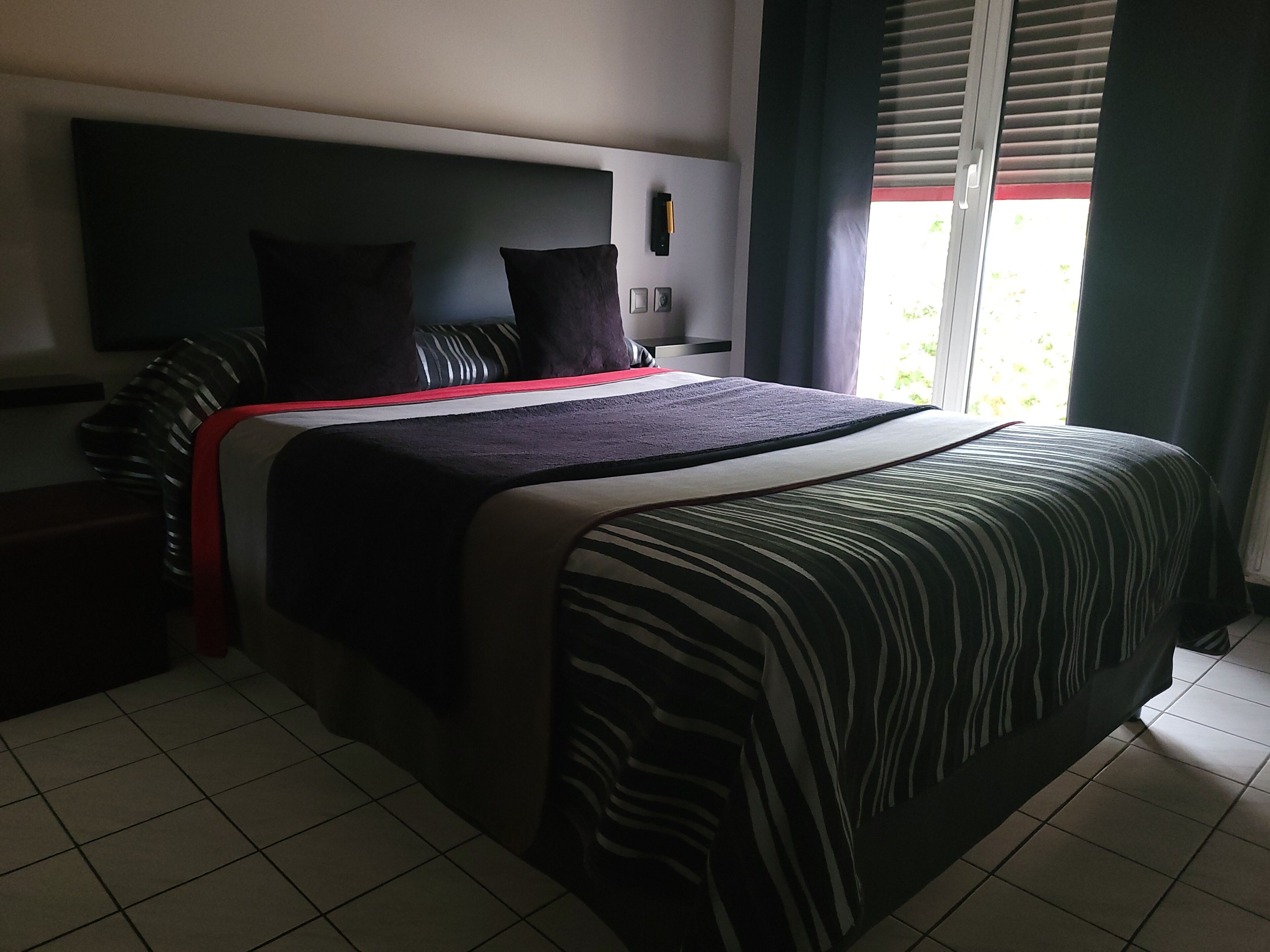 Quarto de hotel moderno com uma cama grande, televisão de tela plana e janela com persianas.