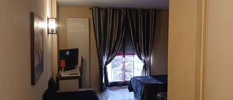 A hotel room with a bed, a desk with a TV, a window with curtains, and a door.