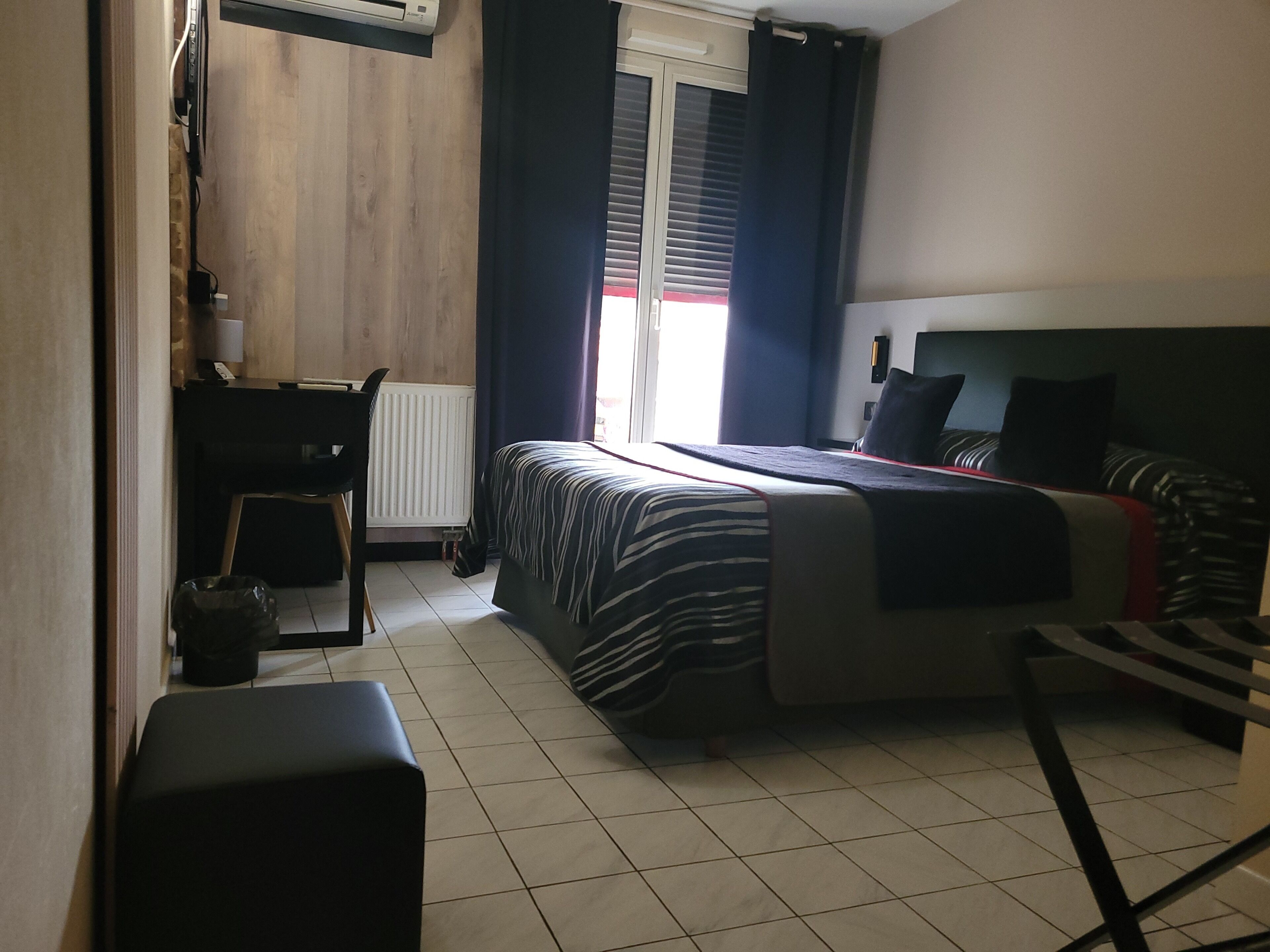 Habitación de hotel con cama, escritorio, silla, radiador y ventana con cortinas.