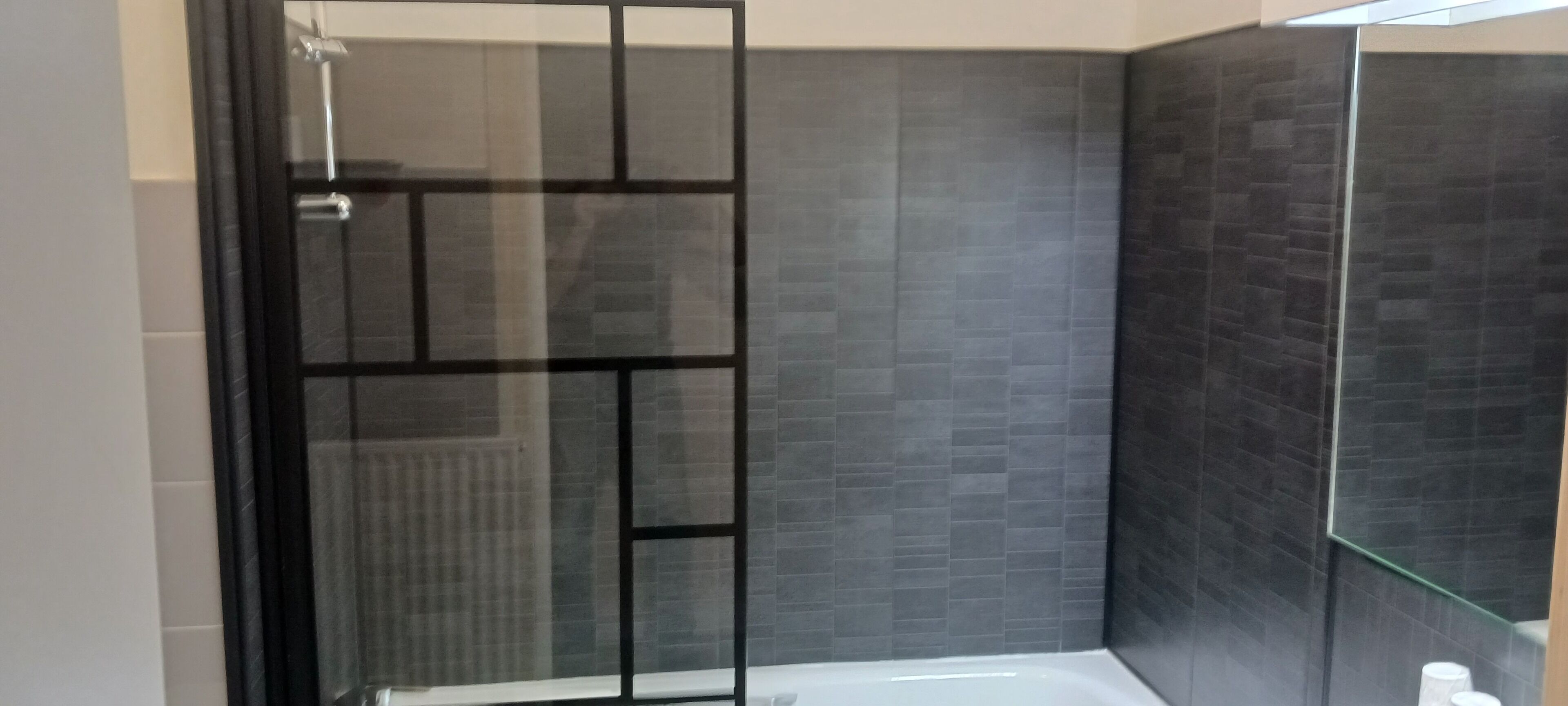 A modern bathroom with a glass shower enclosure, dark tiled walls, and a white bathtub.