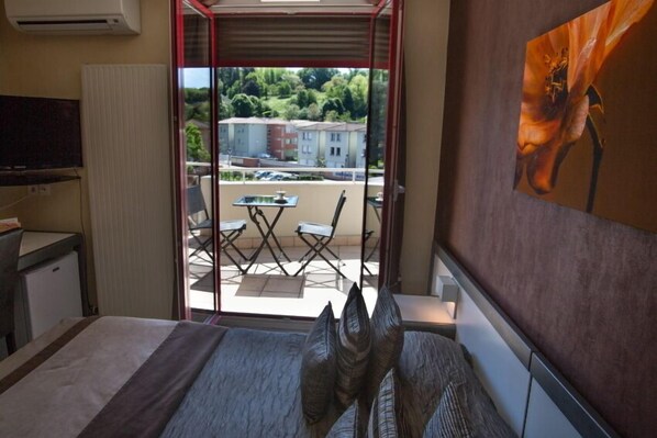 A hotel room with a bed, a TV, a small desk, and a balcony with a table and chairs. - Hôtel Le Médicis (Roussillon-en-Isere)