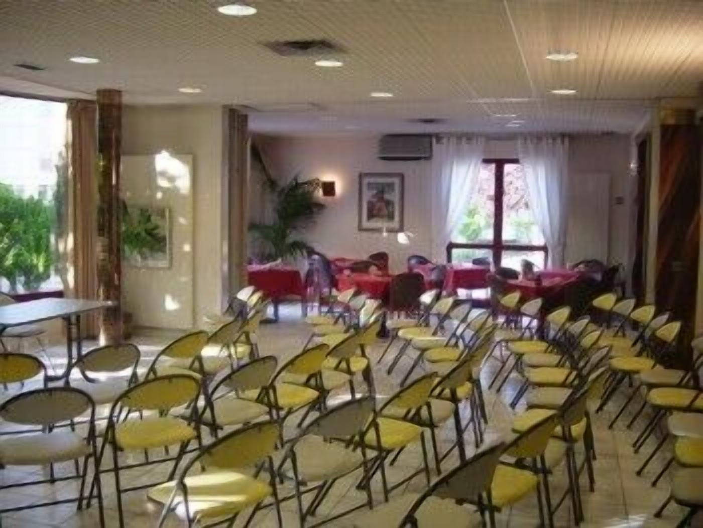A conference room with rows of yellow chairs and tables, large windows with curtains, and indoor plants.
