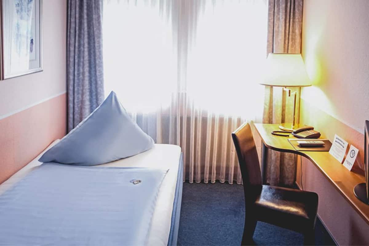 A hotel room with a bed, a desk, a chair, a lamp, and a window with curtains.