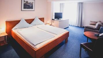 A hotel room with a bed, a desk with a TV, a chair, and a window with curtains.
