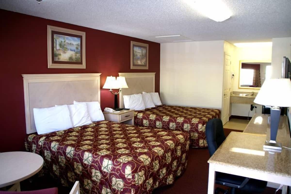 A hotel room with two beds, a desk, and a chair. There are paintings on the walls and a window with curtains.