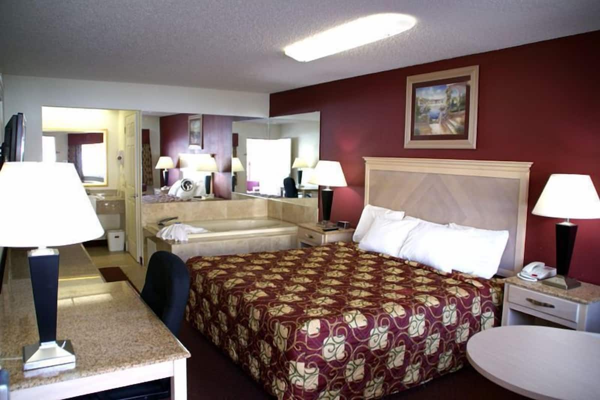 A hotel room with a large bed, two bedside tables with lamps, a round table, a mirror, a TV, and a bathroom with a bathtub and toilet.