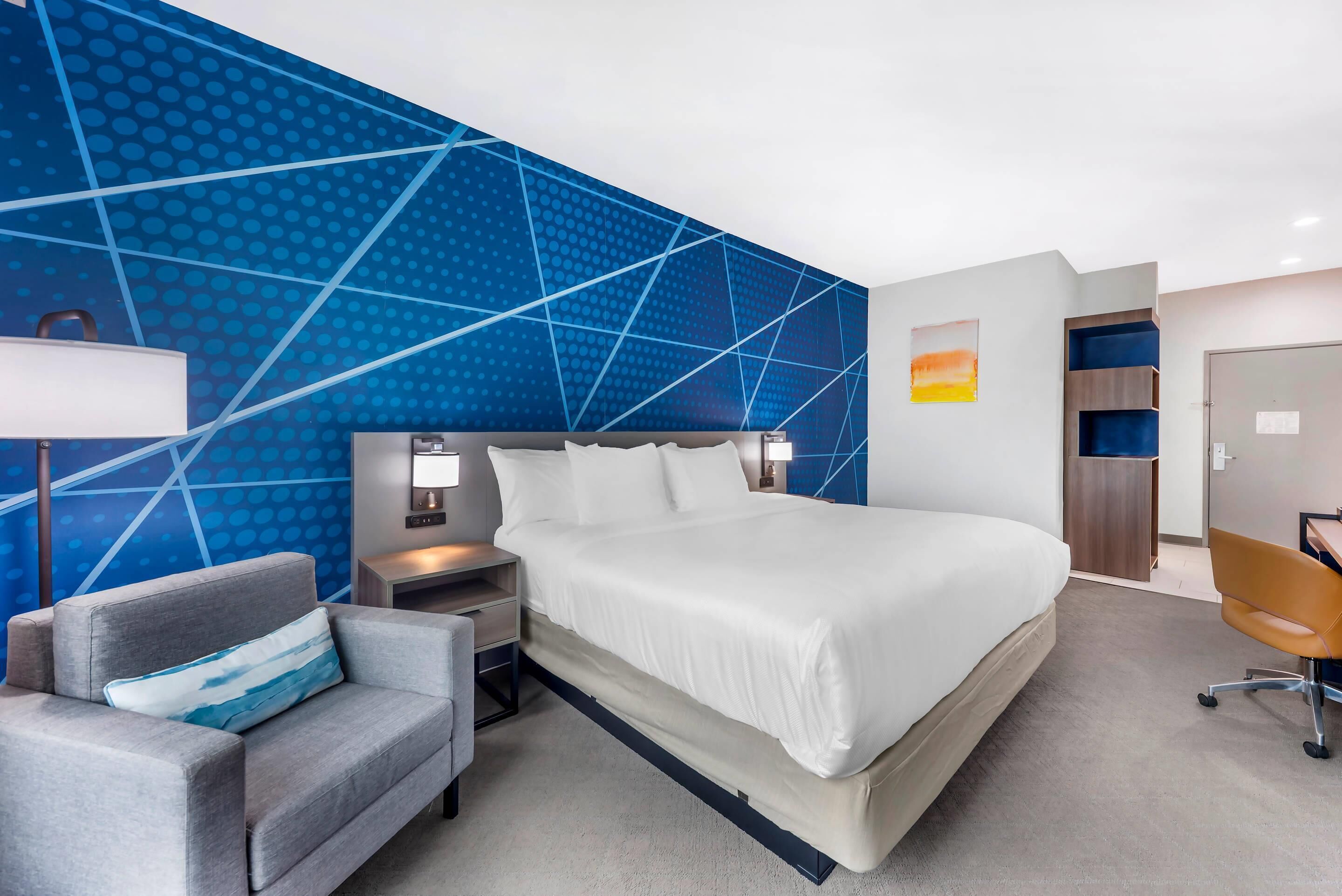 A modern hotel room with a large bed, a gray armchair, a desk, and a blue patterned wall.