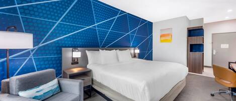A modern hotel room with a large bed, a gray armchair, a desk, and a blue patterned wall.