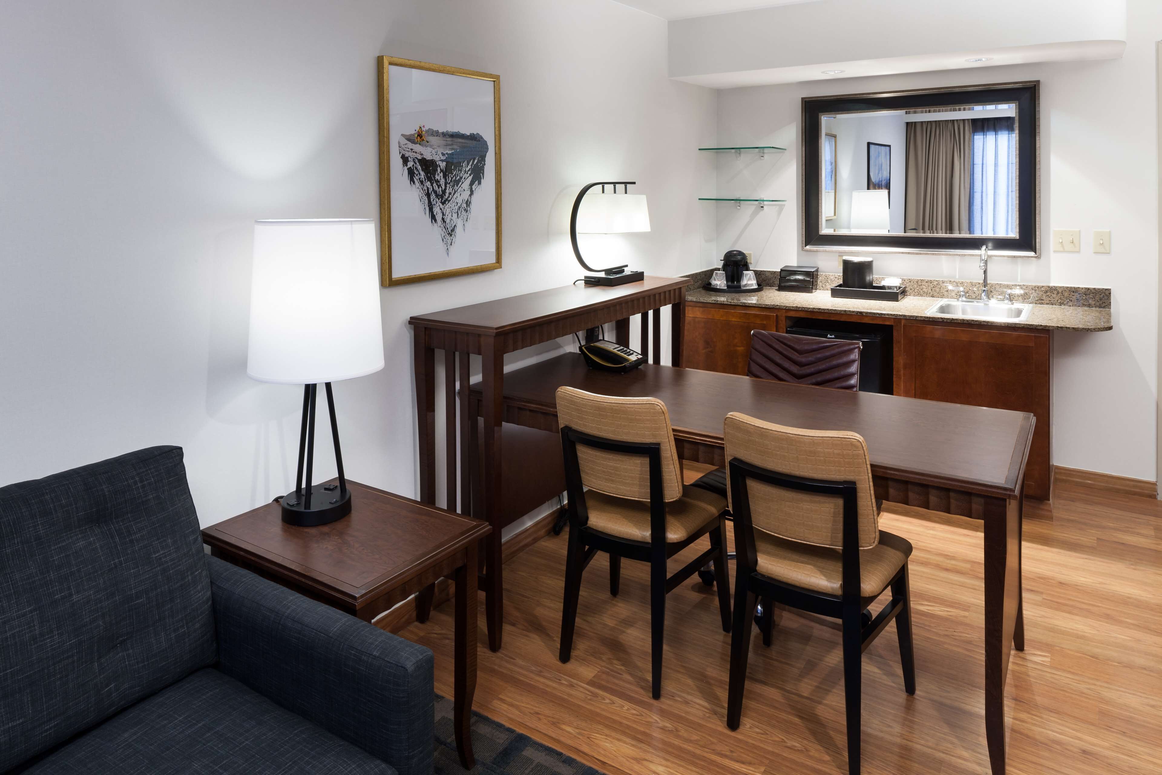 Hotelkamer met een eethoek, een bank, een lamp, een lijst met een afbeelding, een keuken met een spoelbak en een spiegel.