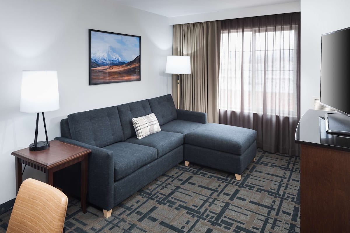 Una habitación de hotel moderna con un sofá modular, una mesita de madera, una silla, un televisor y un ventanal con cortinas.