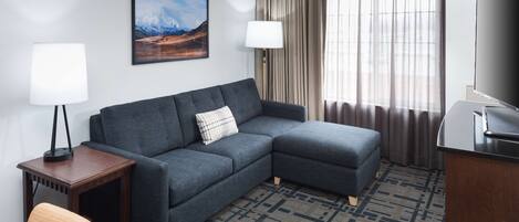 Une chambre d’hôtel moderne avec un canapé d’angle, une petite table d’appoint en bois, un fauteuil, un téléviseur et une grande fenêtre avec des rideaux.