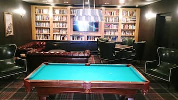 Una mesa de billar en una sala con estantería, sillones de cuero y televisor.