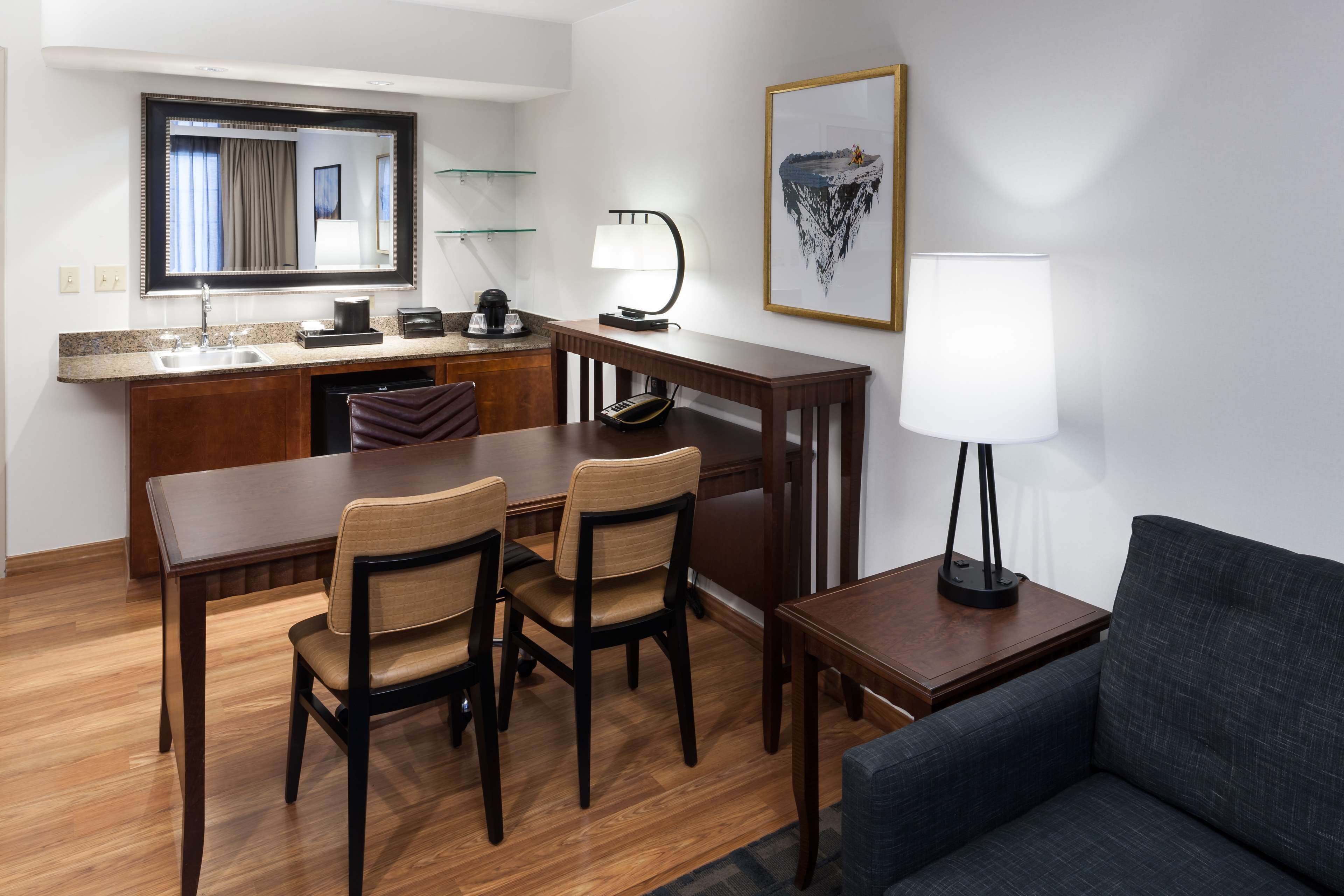A hotel room with a wooden dining table, two chairs, a sofa, a desk with a lamp, a mirror, a framed picture, and a kitchen area with a sink and a coffee maker.
