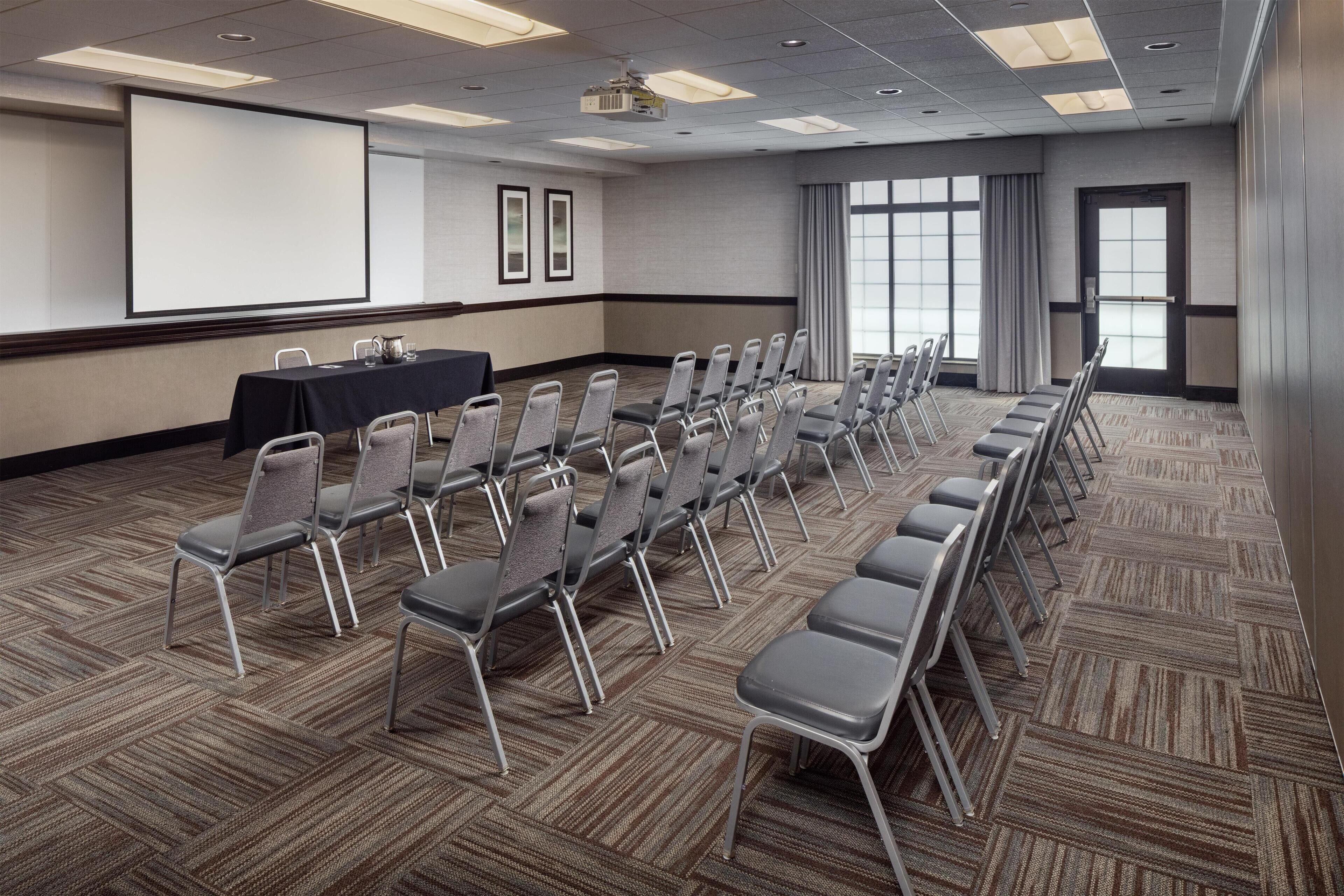 A conference room with rows of chairs, a projector screen, and a table with chairs.