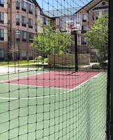 A tennis court with a net, surrounded by a fence, and buildings in the background.