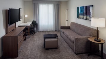 A hotel room with a flat-screen TV, a desk with a chair, a sofa, a small ottoman, a standing lamp, and a painting on the wall.