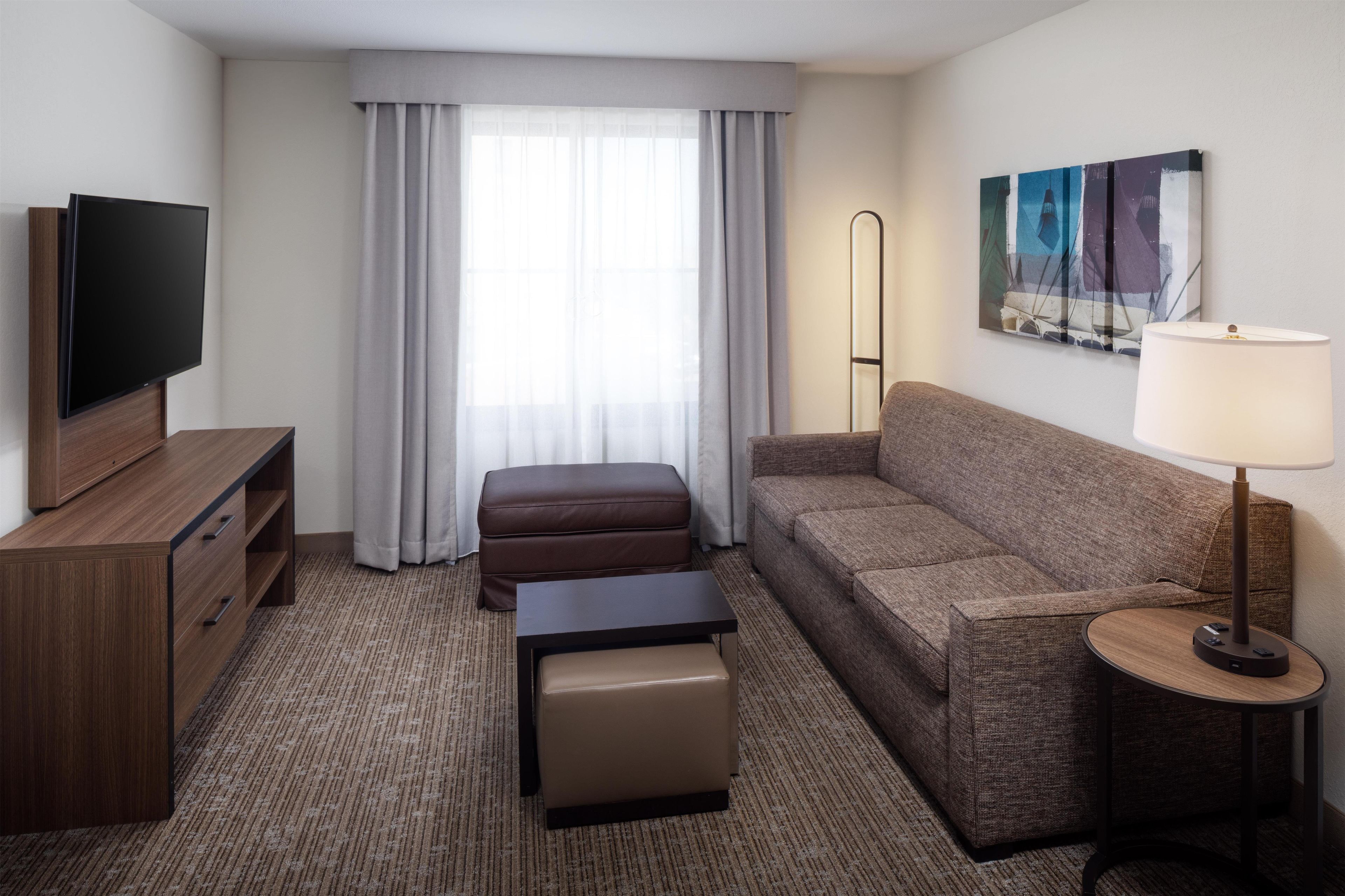 A hotel room with a flat-screen TV, a sofa, a small table, an ottoman, a floor lamp, and a painting on the wall.