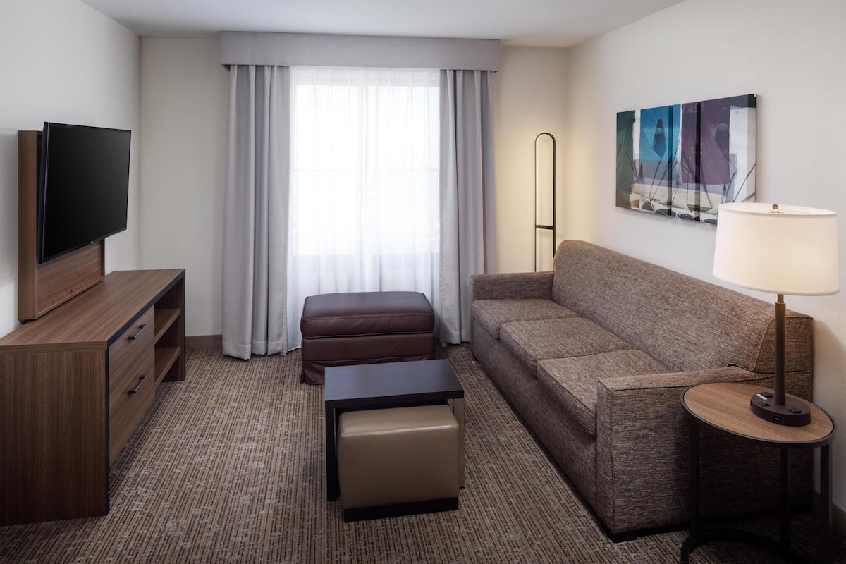 Habitación de hotel con televisor de pantalla plana, sofá, mesita, lámpara, un cuadro en la pared y ventana con cortinas.