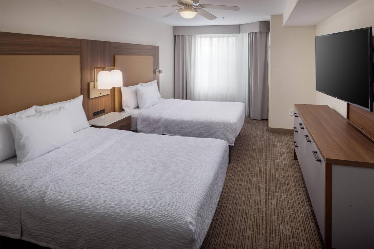 Habitación de hotel con dos camas, televisión, ventilador de techo y ventana con cortinas.