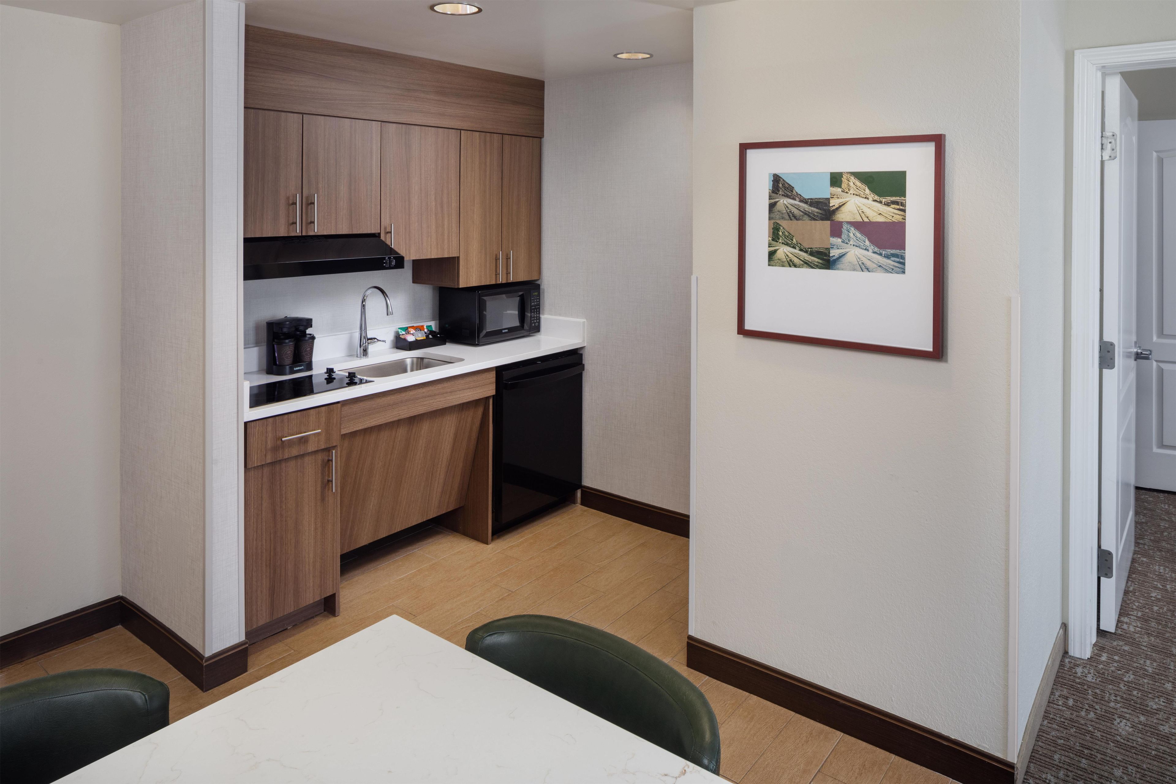 A compact hotel kitchen with a microwave, coffee maker, and a sink. There is a framed picture on the wall and a door leading to another room.