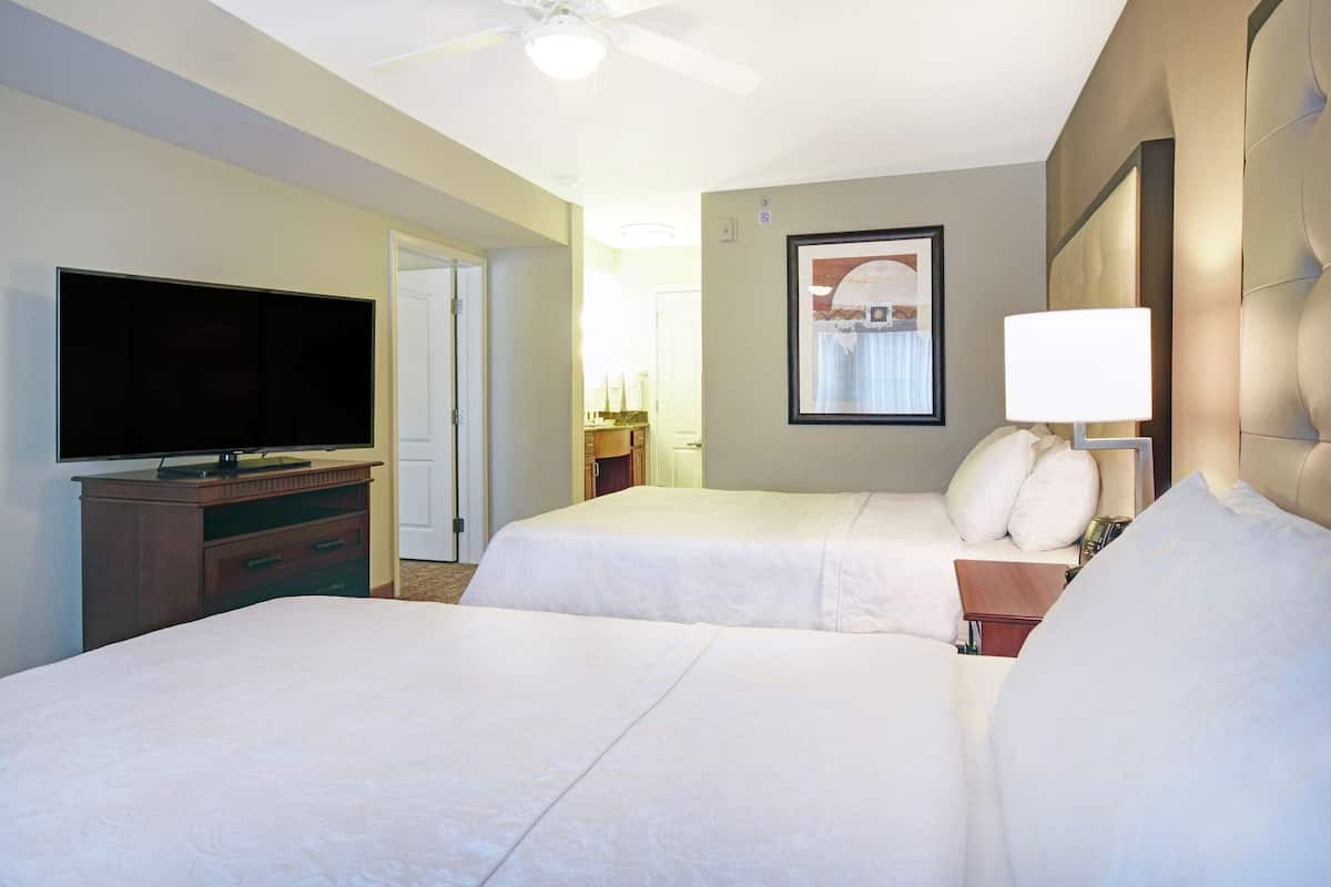 Habitación de hotel con dos camas, un televisor, un ventilador de techo y un cuadro en la pared.
