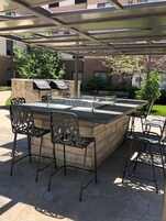 Zona de bar al aire libre con mostrador de piedra, sillas de metal y una parrilla.