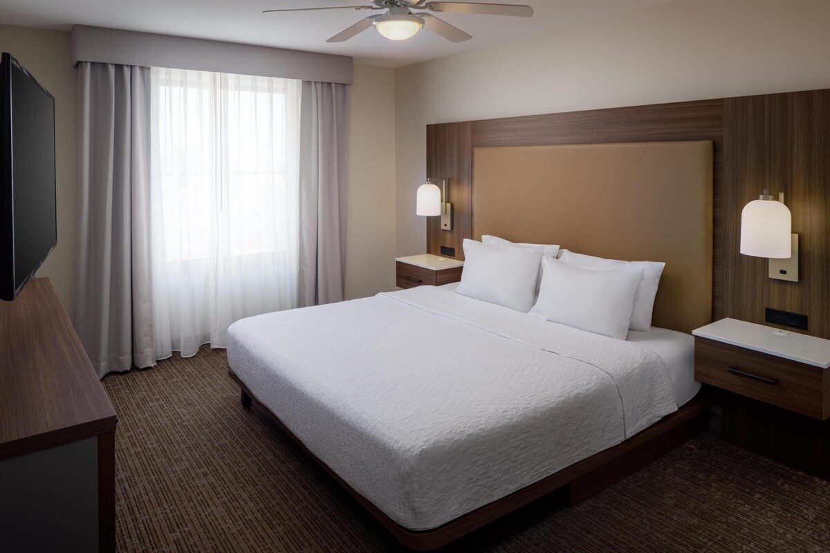 Habitación de hotel con una cama grande, ventilador de techo, televisor de pantalla plana, mesita de noche, lámpara y ventana con cortinas.